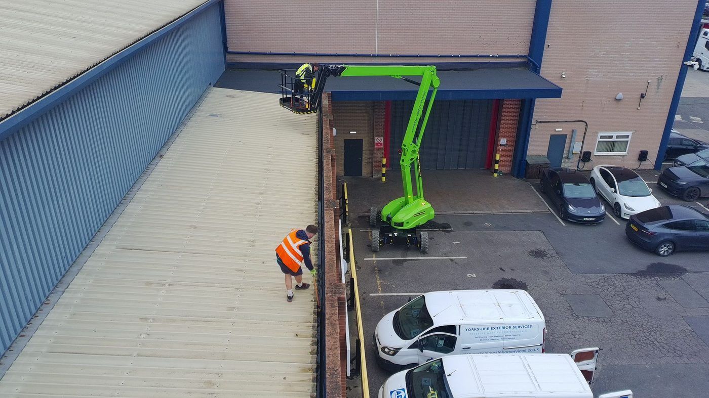 Steel cladding on a commercial unit being washed down, with a green access platform and vans in the yard below