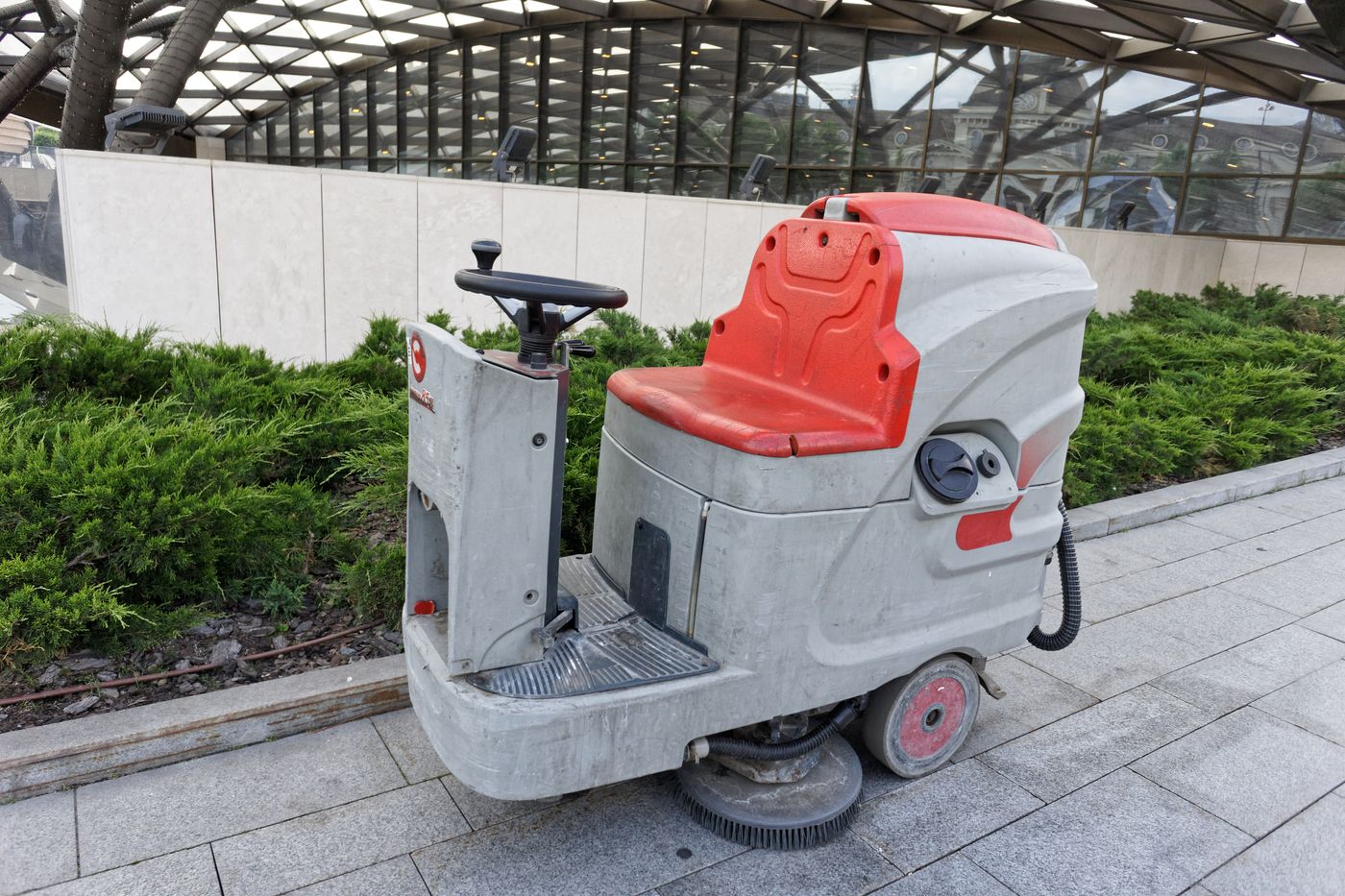 A ride-on scrubber-dryer floor cleaning machine parked on paving beside a planted bed