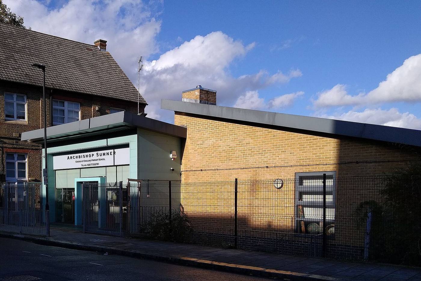 A primary school entrance building in buff brick with railings along the pavement under a blue sky